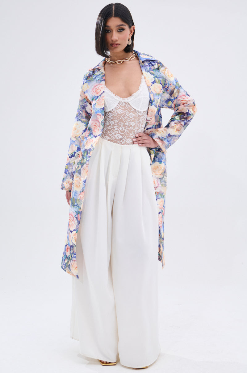 A woman exudes confidence in the PLANT THE SEED SHEER TRENCH, featuring a floral print layered over a white lace bodysuit and wide-legged pants. Her look is completed with straight dark hair, bold makeup, and a chunky gold chain necklace.