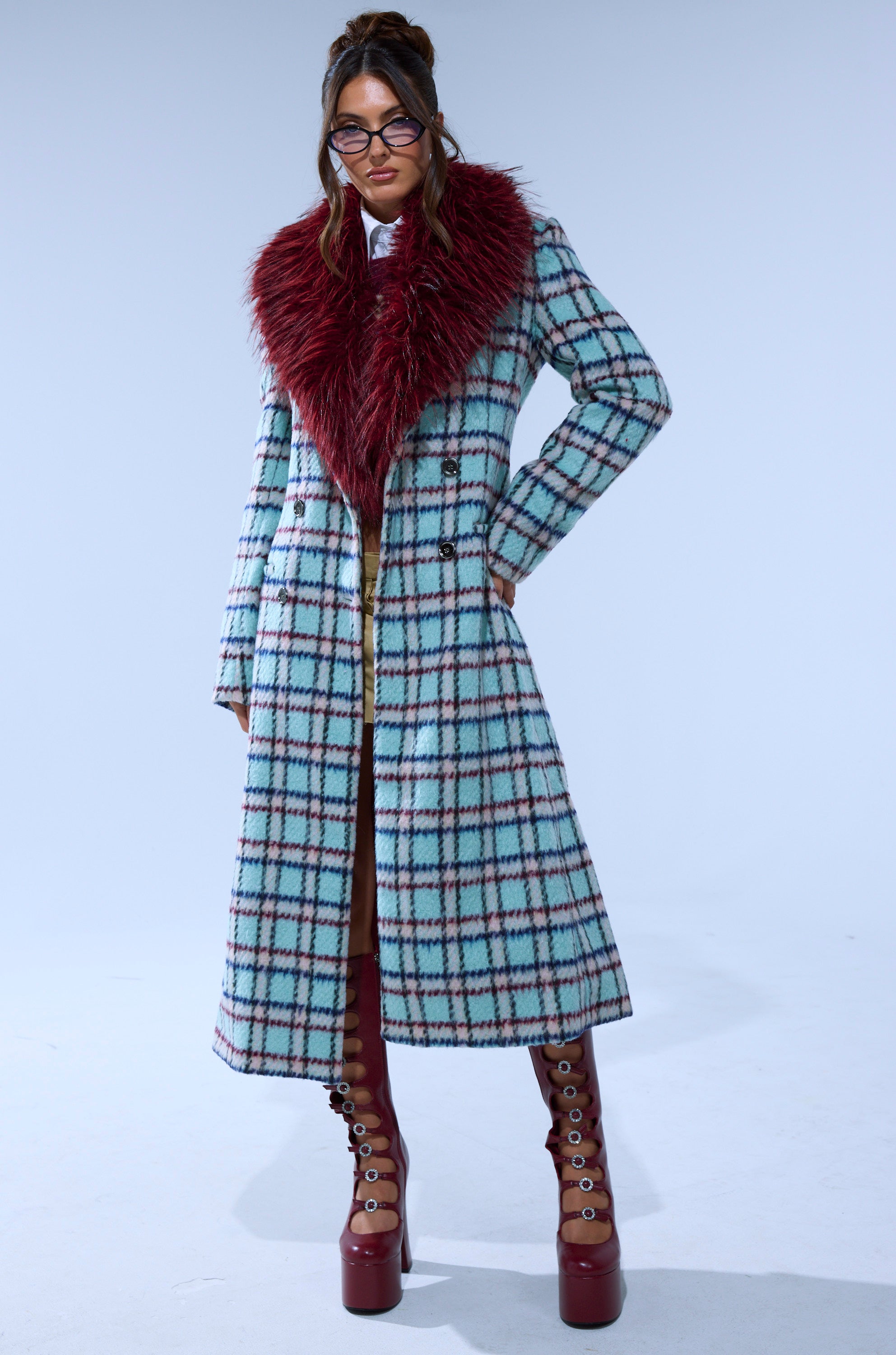 A woman stands confidently against a plain light background, wearing the TEA FOR TWO FUR TRIM TRENCH IN BLUE with a large burgundy faux fur collar, brown knee-high platform lace-up boots, glasses, and styled hair.