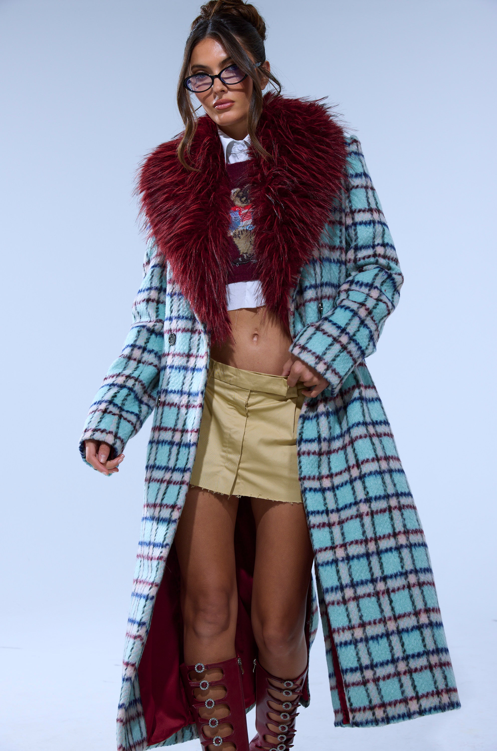 A woman in glasses wears the TEA FOR TWO FUR TRIM TRENCH IN BLUE, paired with a cropped graphic sweater, white shirt, tan mini skirt, and knee-high lace-up boots against a plain background.