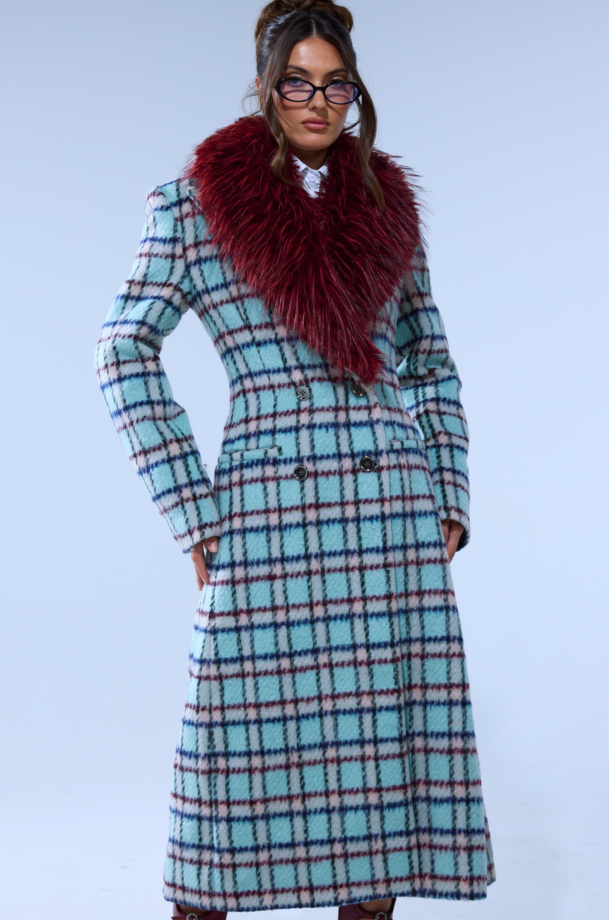 A person wearing the TEA FOR TWO FUR TRIM TRENCH IN BLUE, which features glasses and a large burgundy faux fur collar, stands against a light blue background.