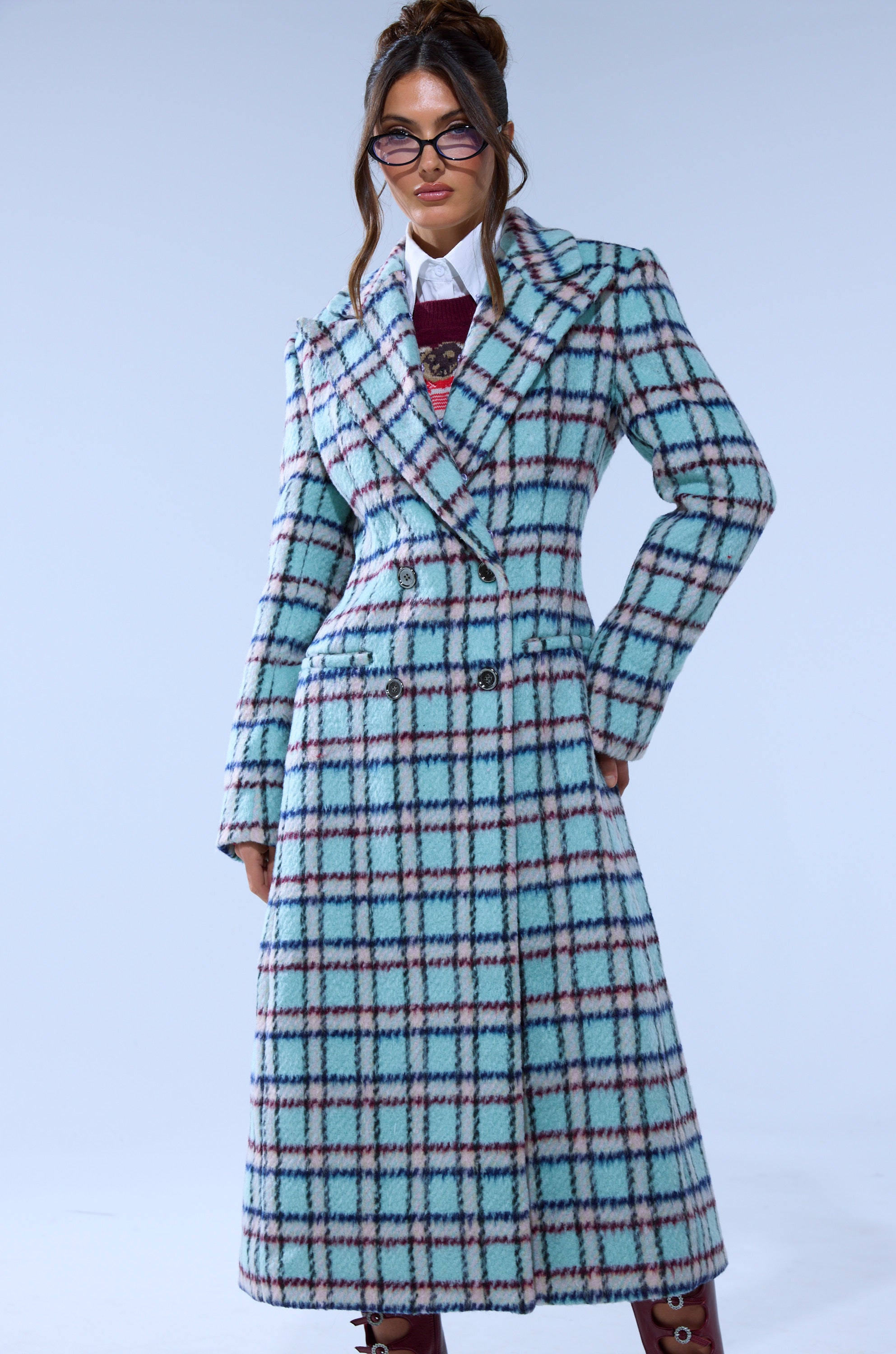 A person poses confidently against a plain light blue background wearing the TEA FOR TWO FUR TRIM TRENCH IN BLUE with a blue plaid pattern featuring pink and black lines, paired with stylish glasses.
