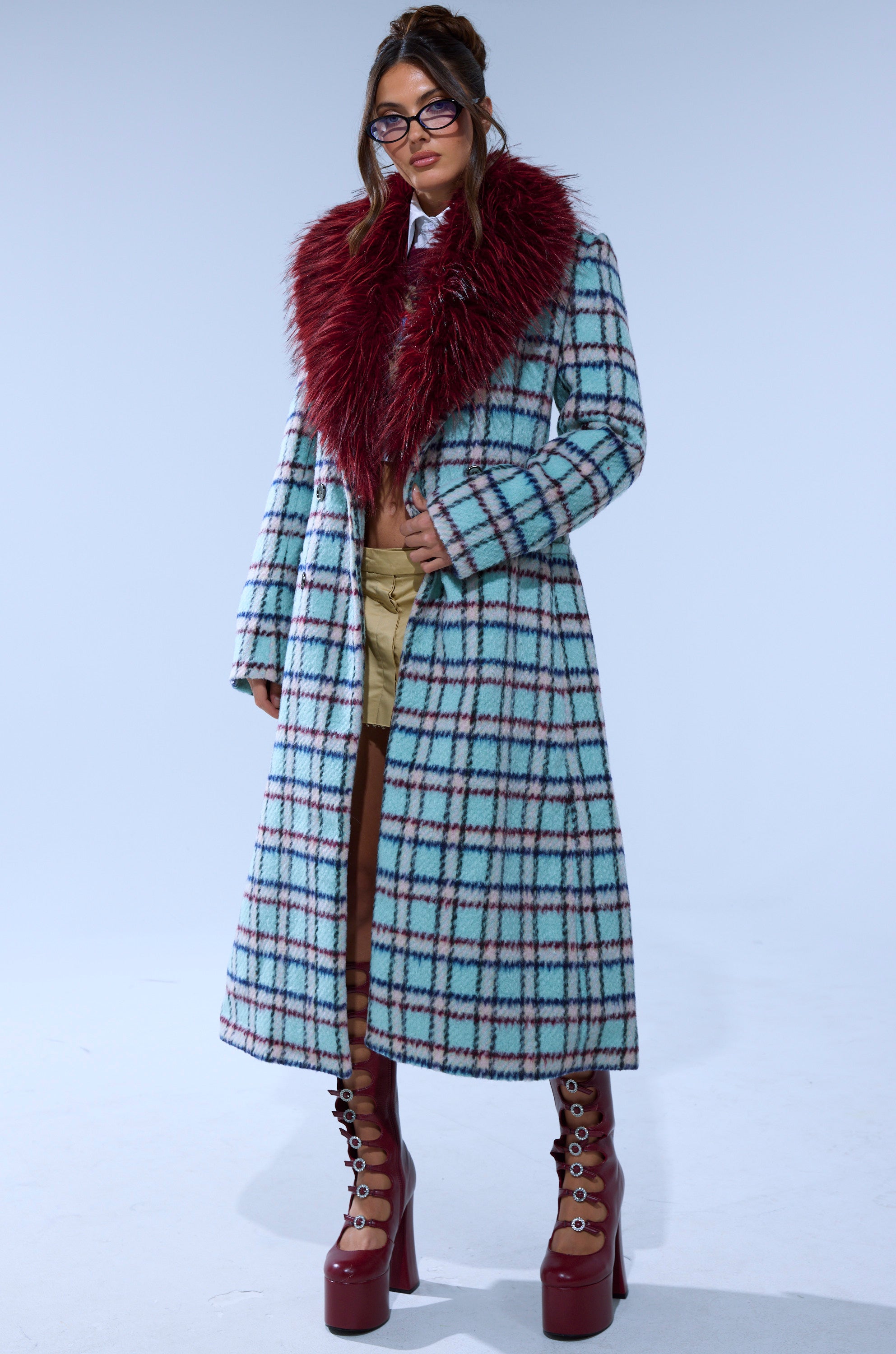 A woman stands against a plain light background wearing the TEA FOR TWO FUR TRIM TRENCH IN BLUE, styled with glasses, tan shorts, tall red lace-up platform boots, and her hair up.