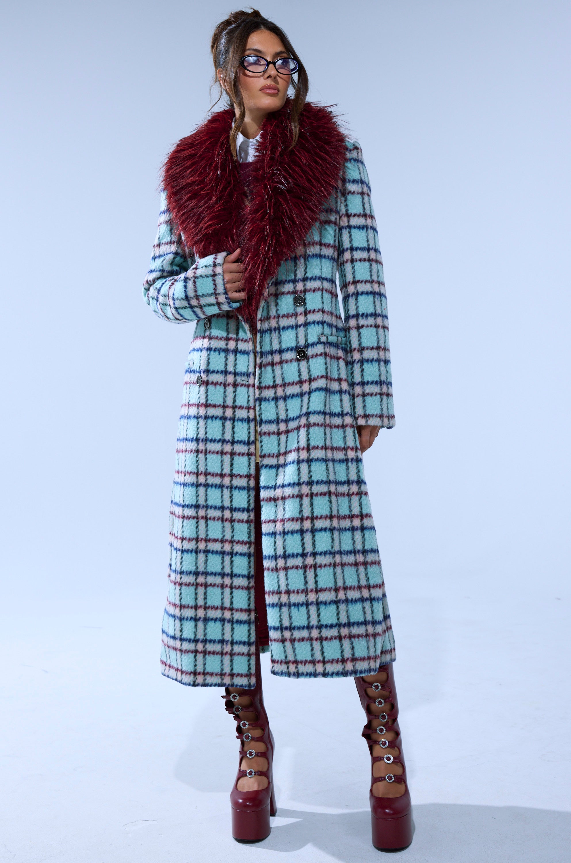 A woman wears the TEA FOR TWO FUR TRIM TRENCH IN BLUE featuring a large burgundy faux fur collar, paired with brown platform boots and glasses, standing against a plain light background.