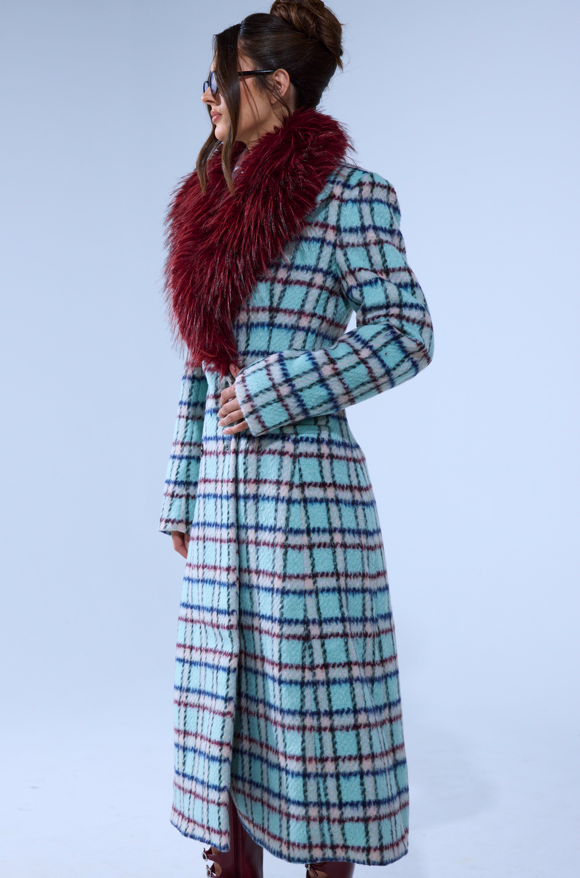 A woman with her hair up and sunglasses stands sideways against a plain background, wearing the TEA FOR TWO FUR TRIM TRENCH IN BLUE—a long blue and white plaid coat with a bold burgundy faux fur collar.