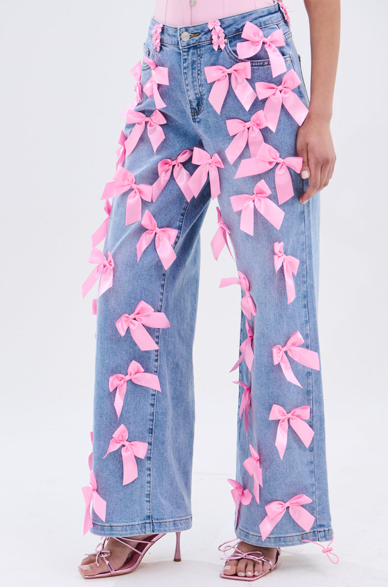 A person wears PRETTY IN PINK BOW DETAIL DENIM JEANS—light blue, wide-leg, high-waisted with pink bow detail—paired with strappy pink heels. The photo is cropped at the shoulders on a plain white background.