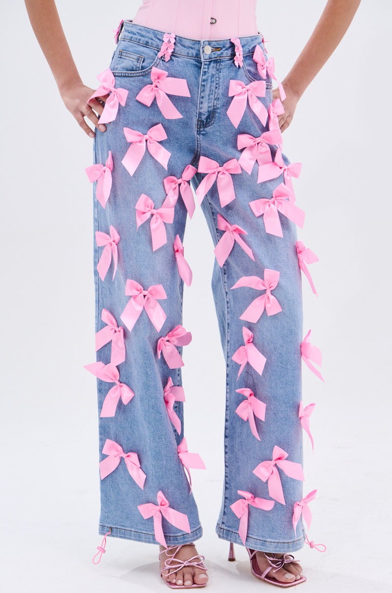 Someone models the PRETTY IN PINK BOW DETAIL DENIM JEANS, featuring high-waisted denim with large pink bows. They pose hands on hips, paired with pink heels and a matching top to show off the bow-adorned jeans.
