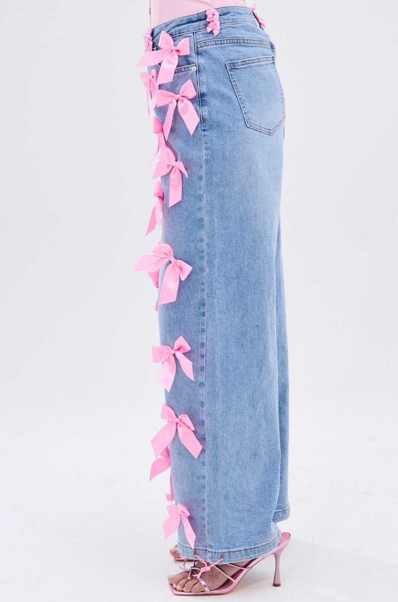 A person wears PRETTY IN PINK BOW DETAIL DENIM JEANS featuring multiple pink bows along the side seam, paired with pink strappy heels, against a plain light background.