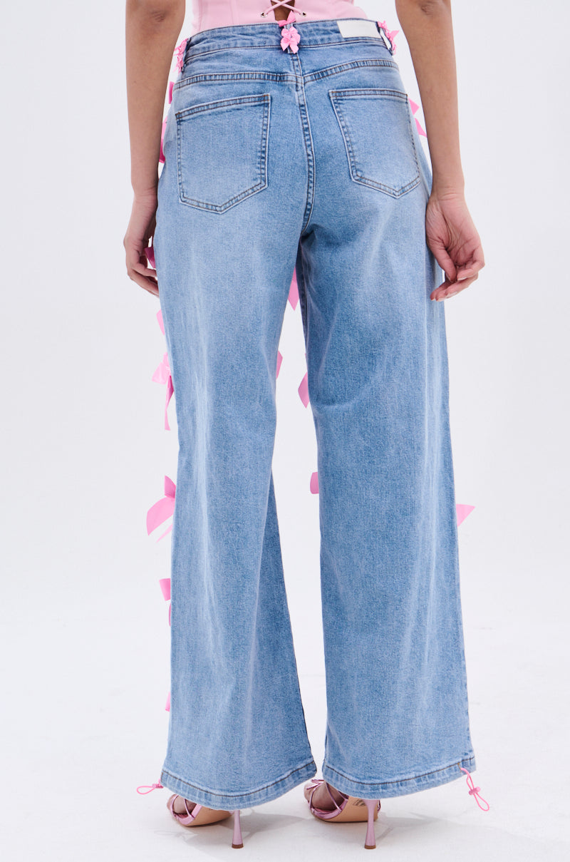 A person wears PRETTY IN PINK BOW DETAIL DENIM JEANS—high-waisted, light blue with pink bows on the sides and back pockets—paired with pink strappy heels against a plain white background.