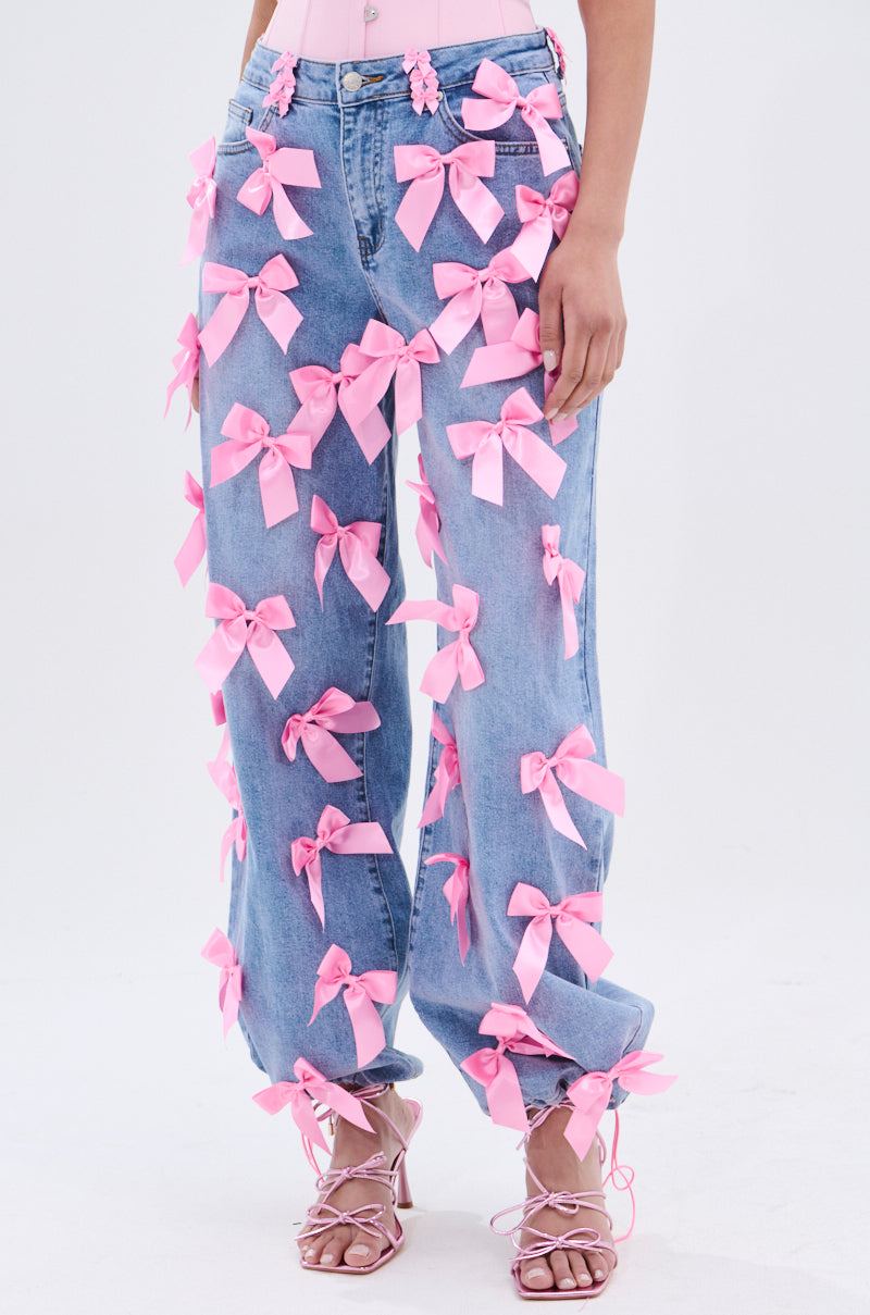 A person models PRETTY IN PINK BOW DETAIL DENIM JEANS—light-wash, high-waisted jeans adorned with pink bows—styled with pink strappy heels. The background is plain white.
