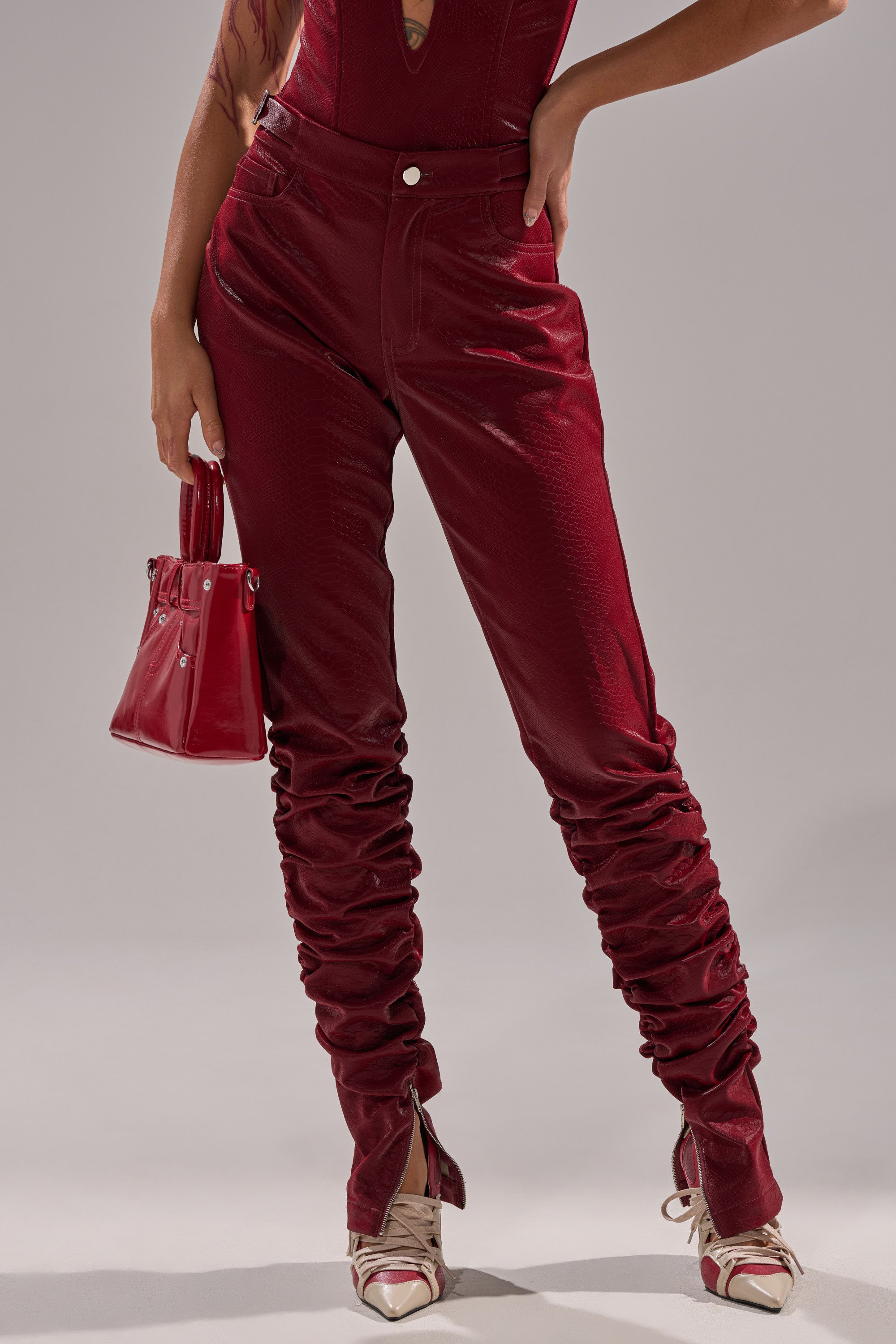 A person wears high-waisted, textured red TOO HOT FOR YOU FAUX LEATHER RUCHED TROUSER, a matching red sleeveless top, red-and-white sneakers, and holds a small red handbag against a plain background.