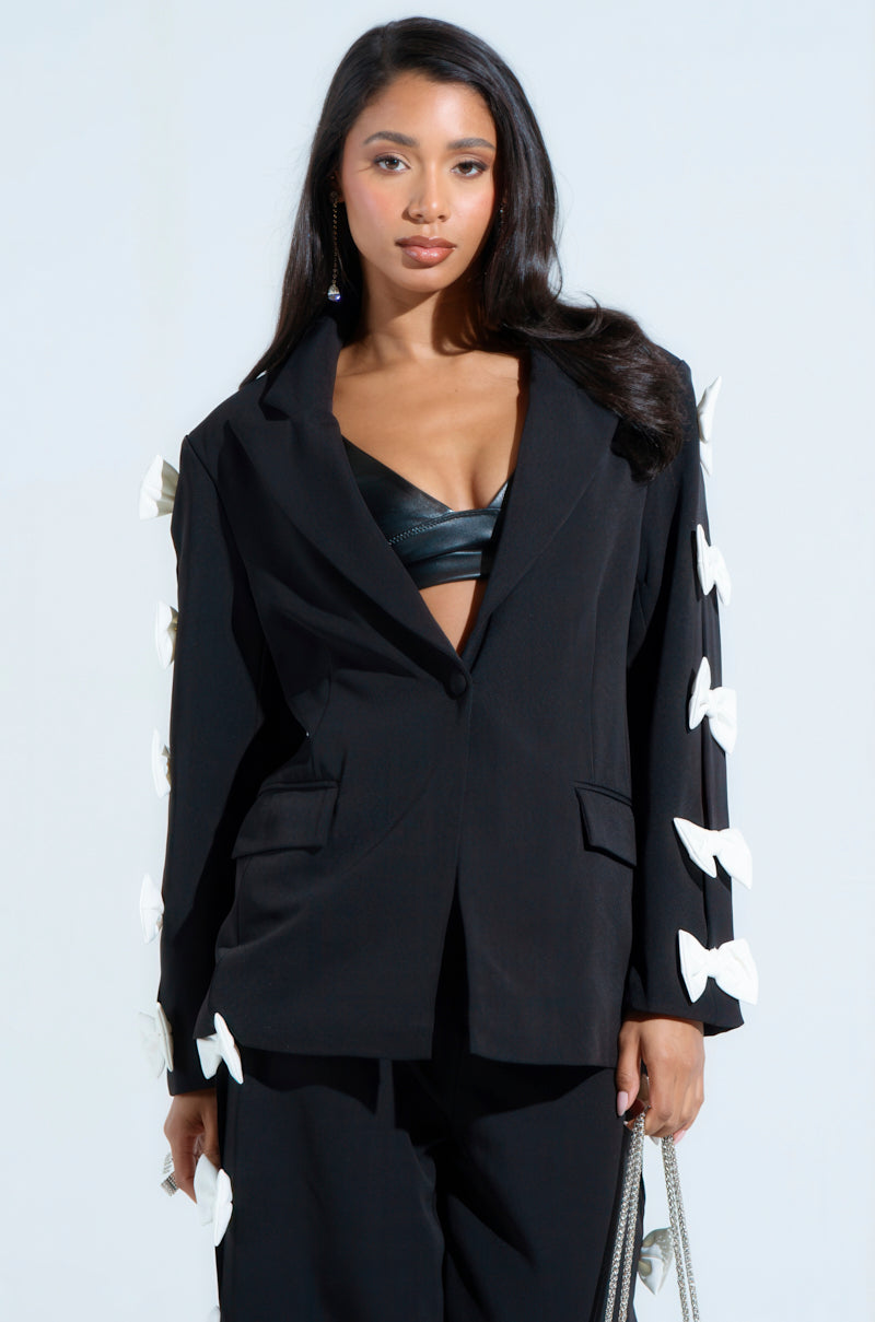 Sporting the FEELING CUTE BOW DETAIL BLAZER, a woman poses with her long, wavy hair cascading and a silver chain purse in hand. Its white accents contrast beautifully against her black top against the light blue background.