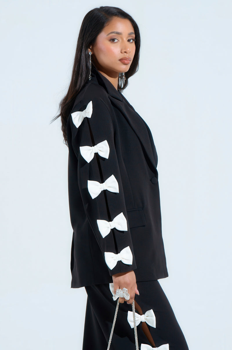 A woman confidently wears the AKIRA Label FEELING CUTE BOW DETAIL BLAZER, showcasing white bows on the sleeves. Her matching black pants follow suit. With long hair and earrings, she holds a silver chain purse against a plain backdrop, truly embracing her stylish look.
