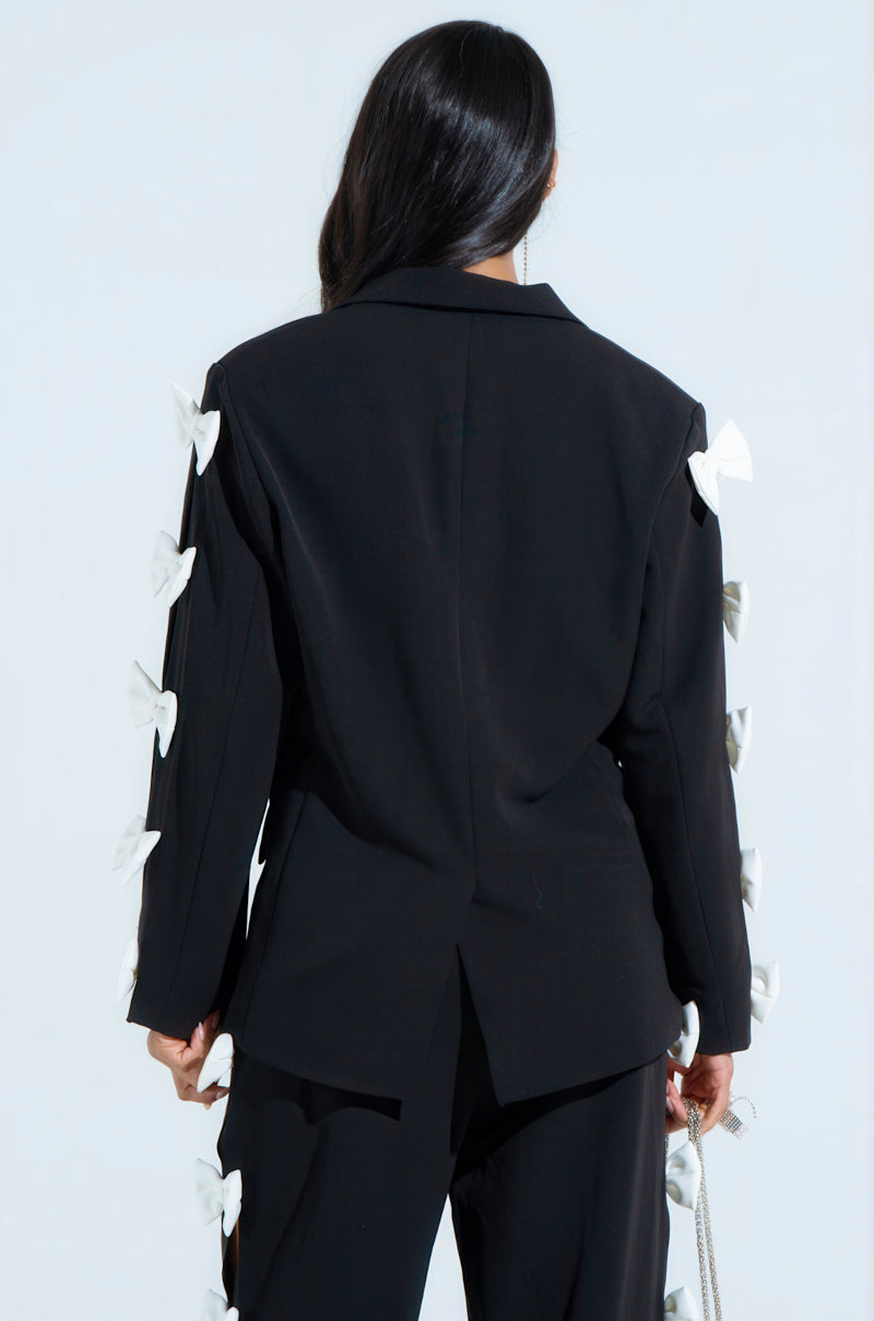 A person with long dark hair in a black FEELING CUTE BOW DETAIL BLAZER, featuring princess seaming and white bow embellishments on the sleeves and pants, stands back to the camera against a light background.
