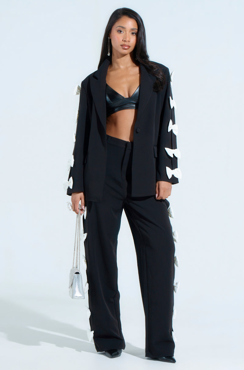 A woman confidently wears the FEELING CUTE BOW DETAIL BLAZER, its princess seaming enhancing her silhouette. She pairs it with a black leather top, a small silver handbag, and long silver earrings against a plain light background.