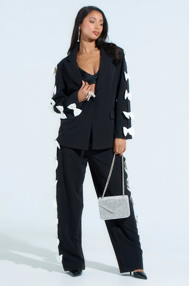 A woman confidently stands in a stylish black pantsuit, spotlighting the "Feeling Cute Bow Detail Blazer" with princess seams and decorative bows along the sleeves and sides. She accessorizes with a sparkly silver handbag and long, dangling earrings, which match her blazer's low-cut top complemented by tonal satiny lining.