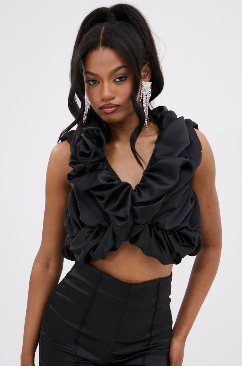 A woman with long, wavy black hair and rhinestone earrings layers the SOMETHING SWEET SATIN BOLERO over a sleeveless black top and high-waisted pants, creating a high-fashion look against a plain light background.