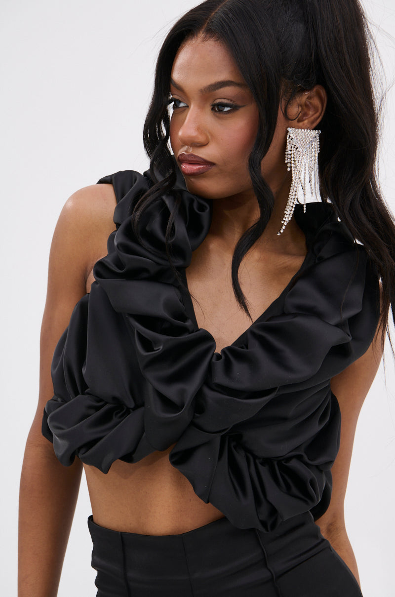 Wearing the SOMETHING SWEET SATIN BOLERO and dramatic dangling pearl earrings, a woman with long, wavy black hair poses elegantly with a serious gaze, her chic look enhanced by the bolero against a plain background.
