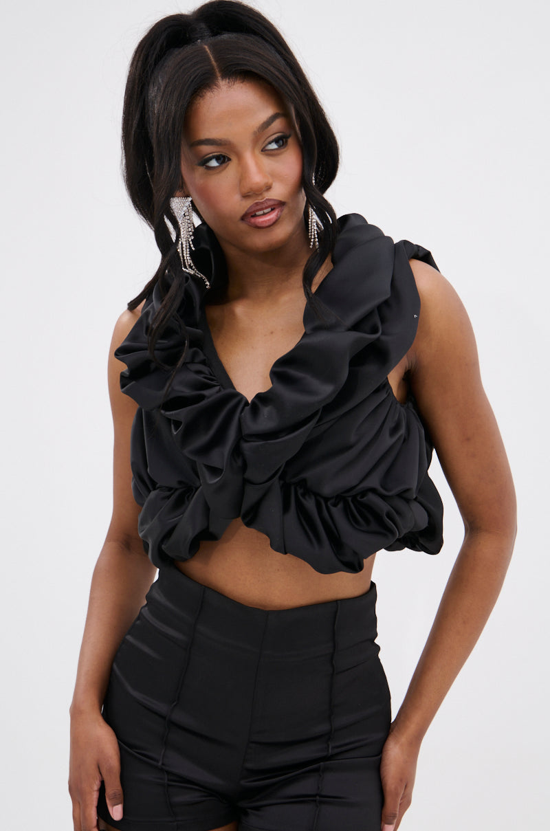 A woman with long dark hair wears the SOMETHING SWEET SATIN BOLERO over black high-waisted shorts, accessorized with large statement earrings. She looks slightly left against a plain background for a chic, high-fashion vibe.