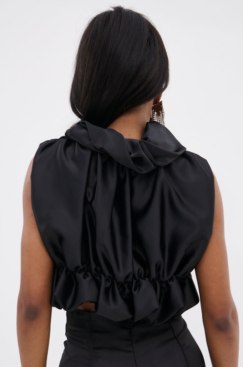 A person with long, straight dark hair is seen from behind wearing the SOMETHING SWEET SATIN BOLERO featuring oversized ruffles and a gathered back. They pair it with large, dangling earrings.