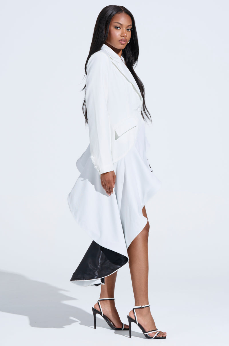 A woman in a white ensemble, highlighted by the MAKING PLANS ASYMMETRICAL BLAZER and a flowing asymmetrical skirt, poses against a plain backdrop. Her strappy high heels complement her cascading long dark hair and exude tailored sophistication and confidence.