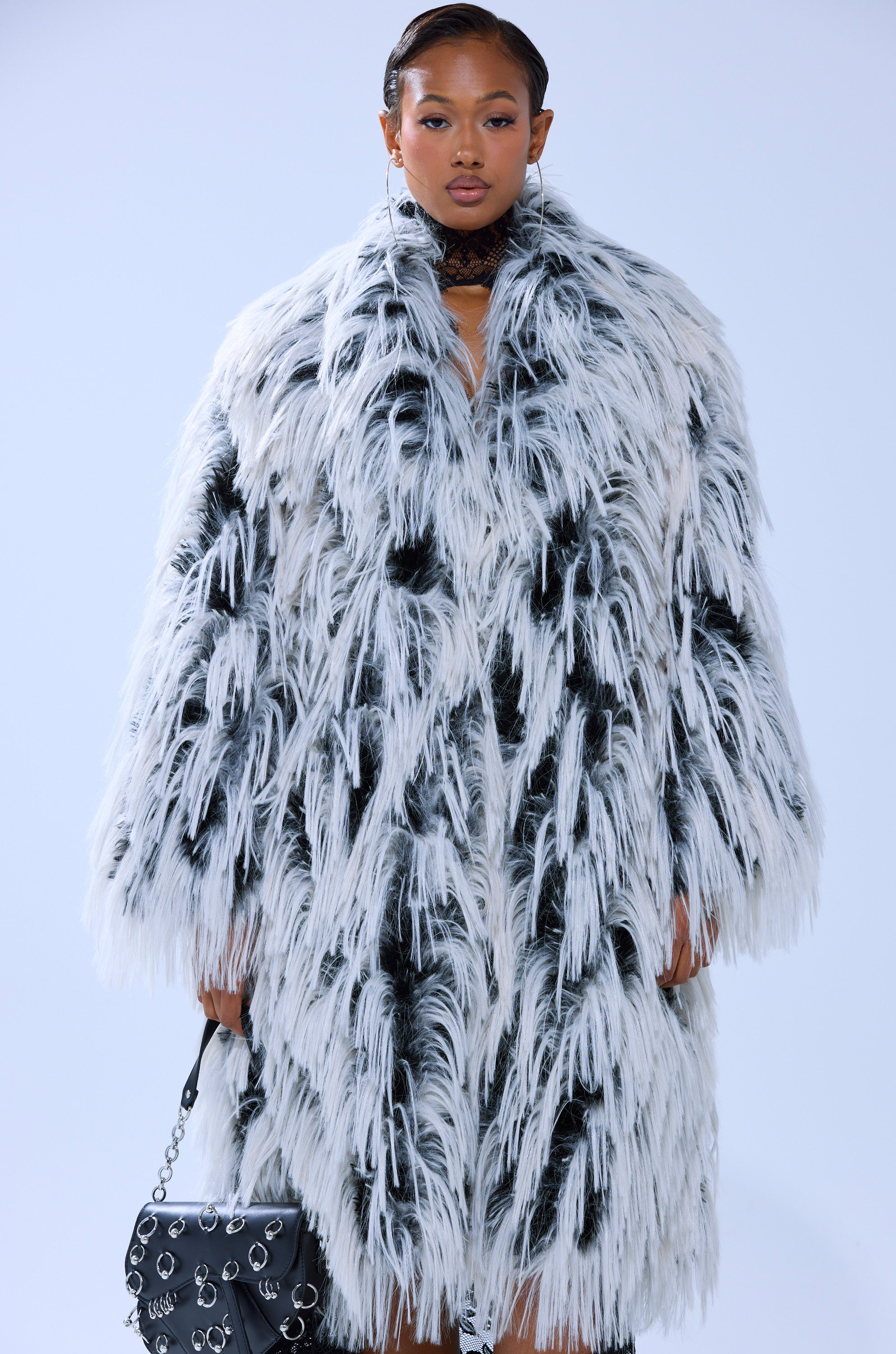 A model wears the EXPENSIVE TASTE FAUX FUR coat—an oversized, longline black and white shaggy faux fur piece—paired with a black purse featuring silver chain and ring accents, against a plain light background.
