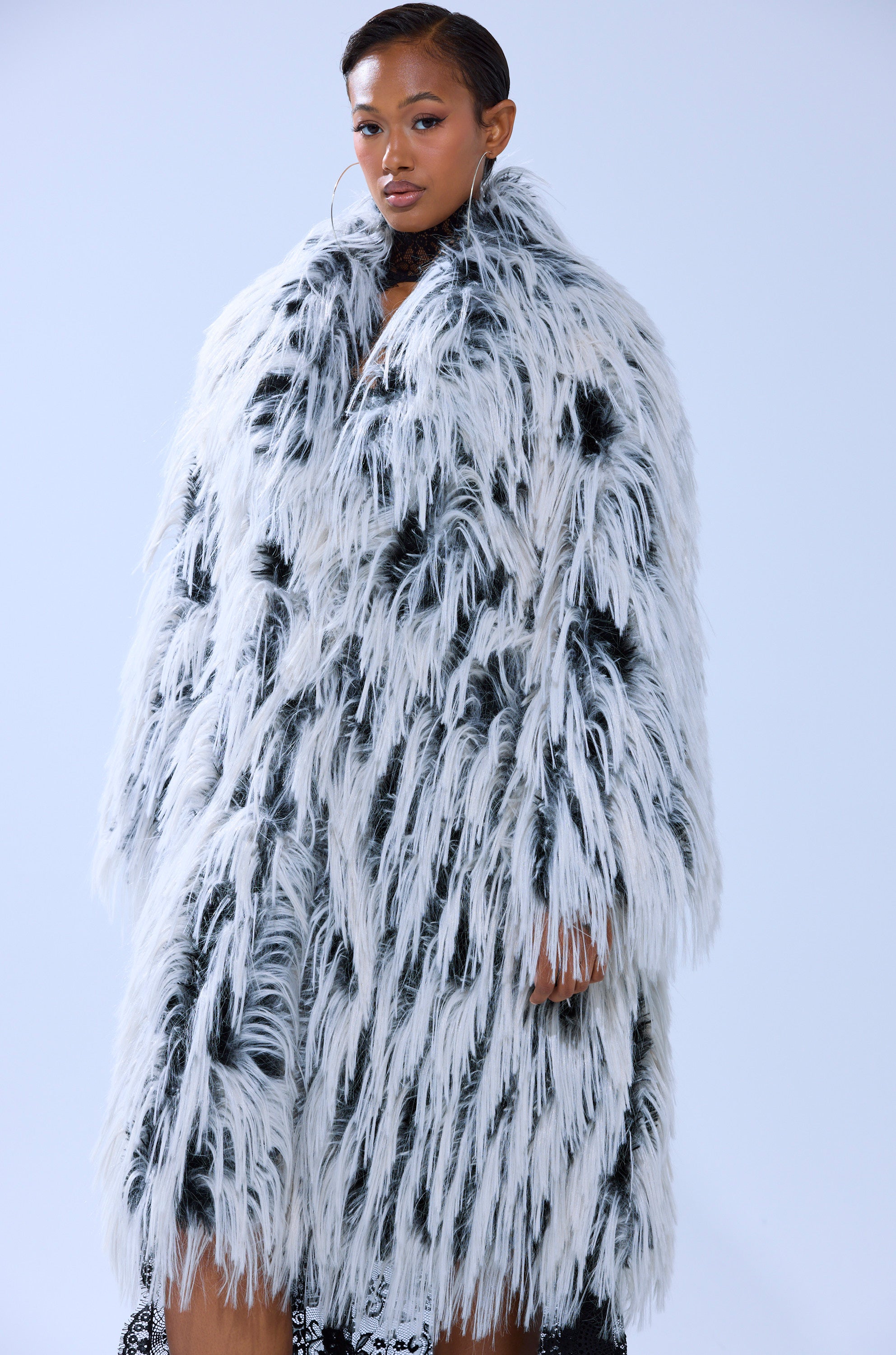 A woman stands confidently in the EXPENSIVE TASTE FAUX FUR, a bold, oversized longline jacket with black and white shaggy texture, making a striking statement against a light background.