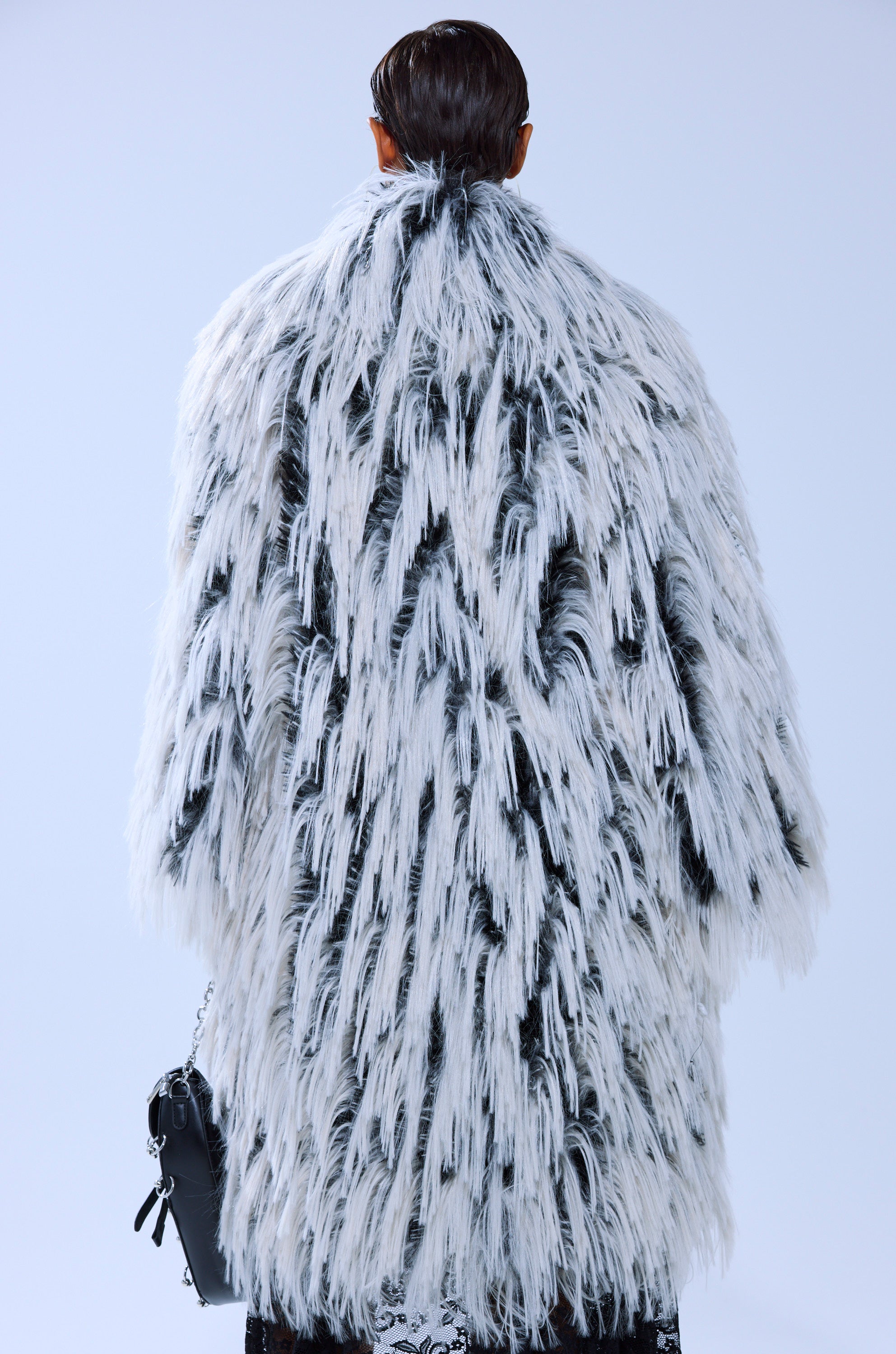 A person with slicked-back hair is seen from behind, wearing the EXPENSIVE TASTE FAUX FUR longline black and white coat and holding a small black handbag against a light background.