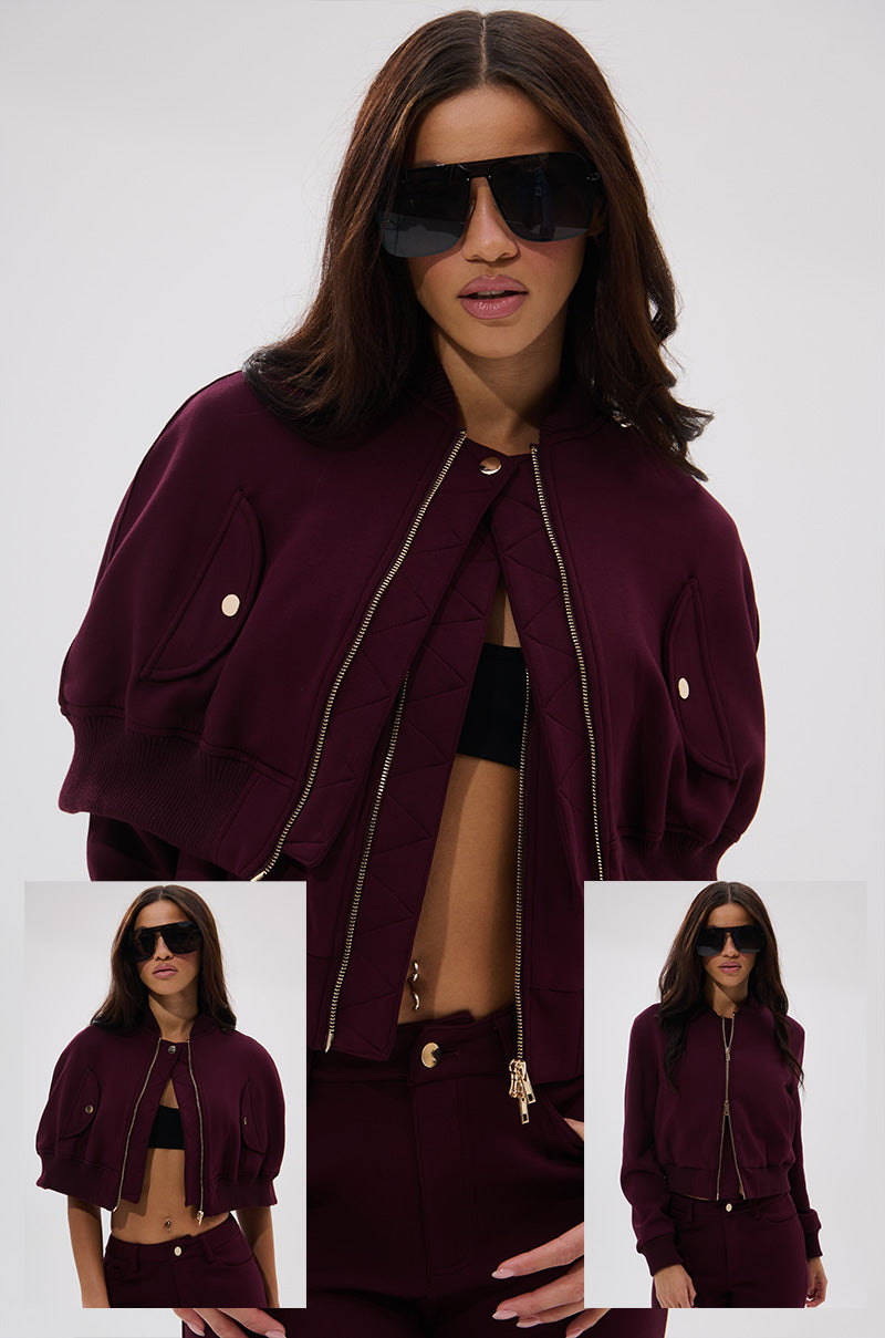 A woman models the LYNX LAYERED BOMBER JACKET IN BURGUNDY with matching pants, a black top, and bold sunglasses. Two inset images highlight the jacket’s double-layer design from different angles.