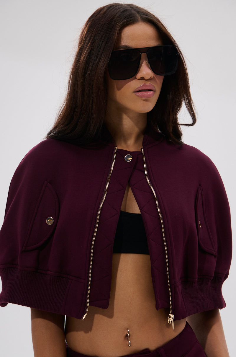 A woman with long straight brown hair, dark sunglasses, and a belly button piercing wears the LYNX LAYERED BOMBER JACKET IN BURGUNDY over a black bandeau top and maroon pants. The background is plain light gray.