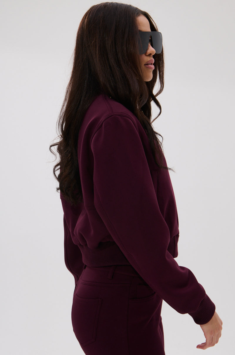 Wearing oversized black sunglasses and the LYNX LAYERED BOMBER JACKET IN BURGUNDY with matching pants, a person with long, wavy dark hair stands sideways against a light gray background.