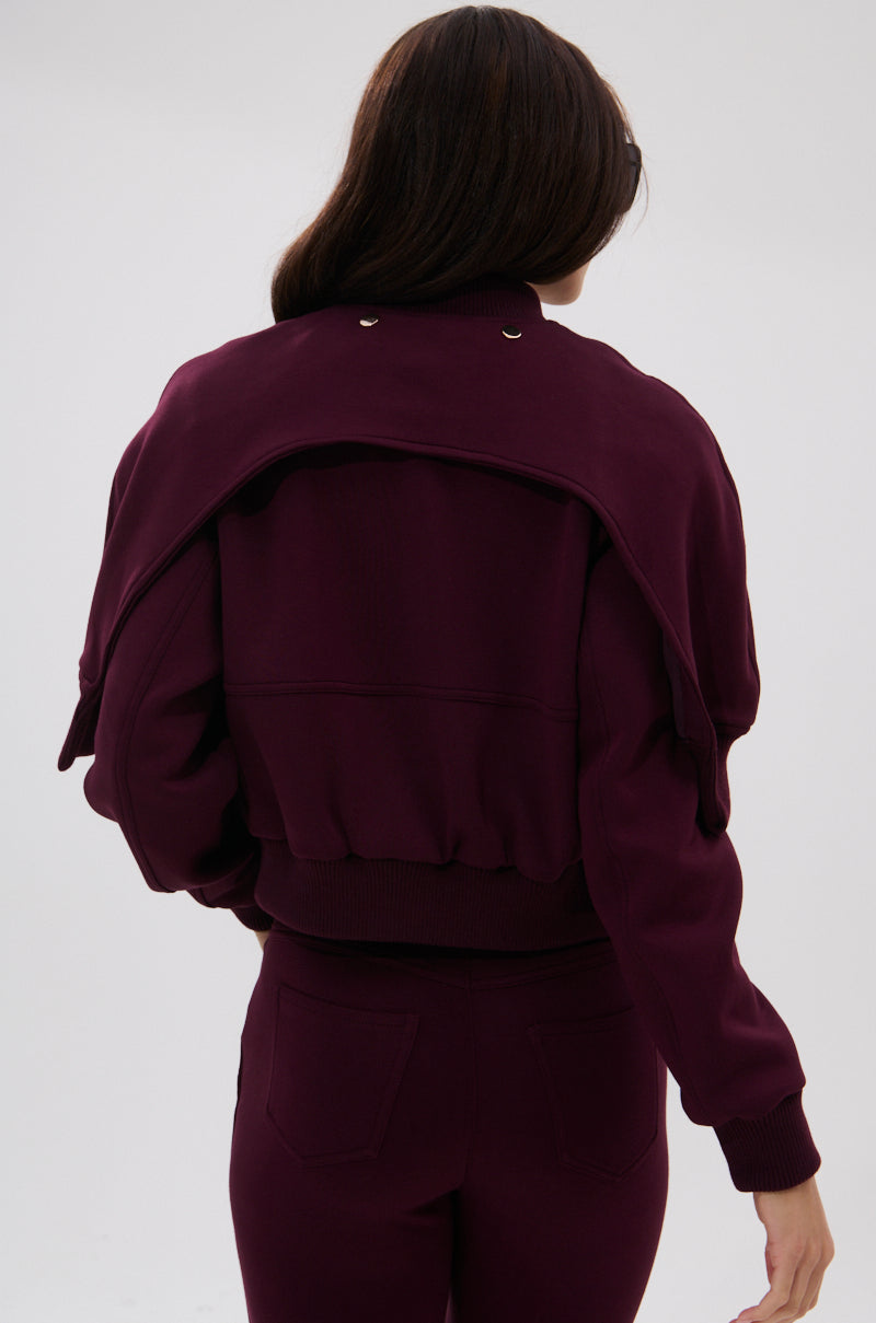 A woman with long brown hair, seen from behind, wears a dark purple sweatshirt and pants, with the sweatshirt draped over her shoulders to reveal the LYNX LAYERED BOMBER JACKET IN BURGUNDY underneath.