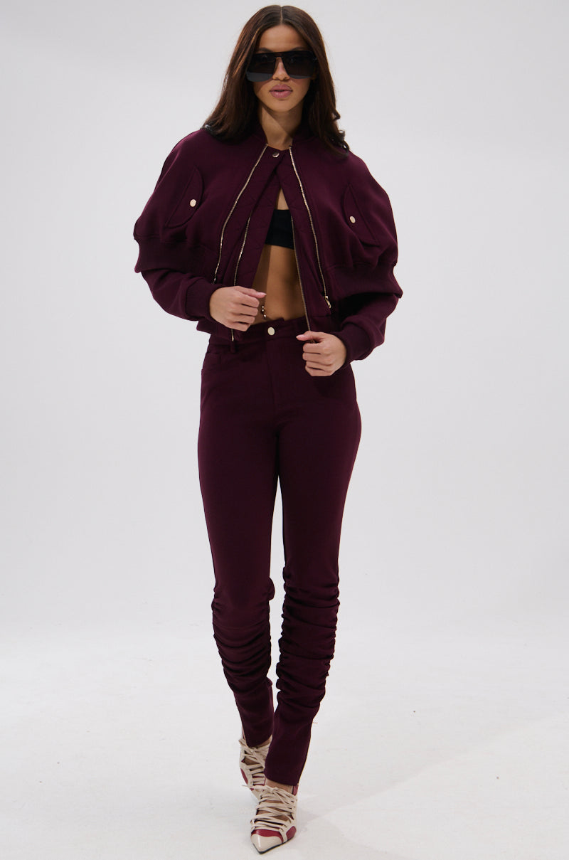 A woman in the LYNX Layered Bomber Jacket in Burgundy, paired with high-waisted ruched pants, a black bralette, sunglasses, and strappy heels, stands against a plain white background.