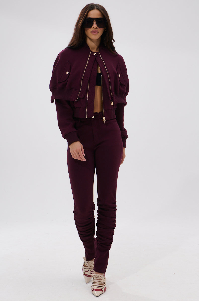A woman wearing the LYNX LAYERED BOMBER JACKET IN BURGUNDY with matching ruched pants, a black bralette, strappy heels, and sunglasses walks confidently against a plain white background.