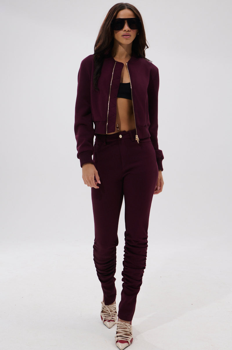 A woman in sunglasses wears the LYNX LAYERED BOMBER JACKET IN BURGUNDY with matching high-waisted pants, a black crop top, and striped pointed shoes, standing against a plain white background.