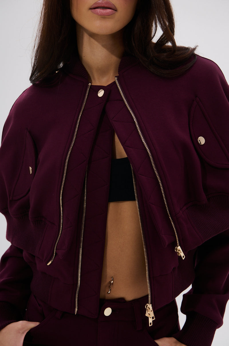 A woman wears the LYNX Layered Bomber Jacket in Burgundy, featuring gold zippers and buttons, partially unzipped to show a black crop top and high-waisted pants. Only her lower face is visible.