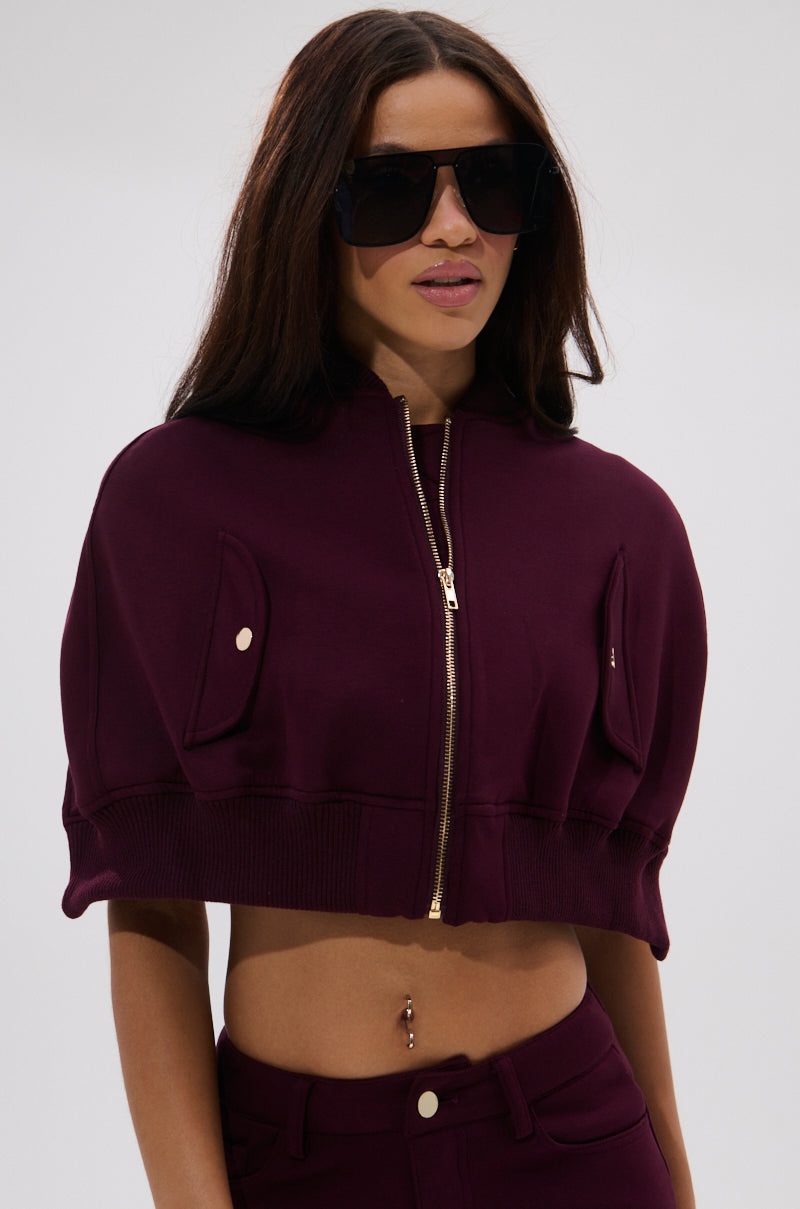 A woman wears the LYNX LAYERED BOMBER JACKET IN BURGUNDY, styled as a sleek two-piece set with oversized black sunglasses, standing against a plain light background.