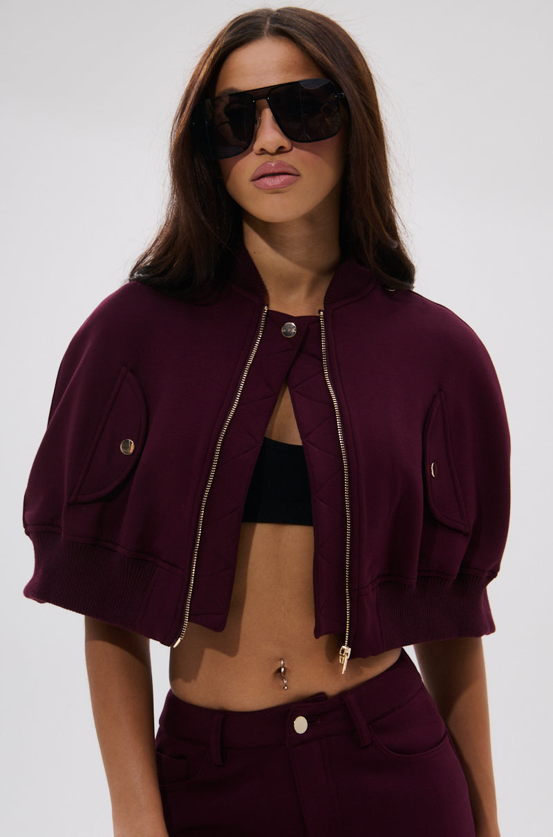 A woman in the LYNX LAYERED BOMBER JACKET IN BURGUNDY with gold zippers, large black sunglasses, a black bandeau top, maroon pants, and a belly button piercing stands against a plain light background.