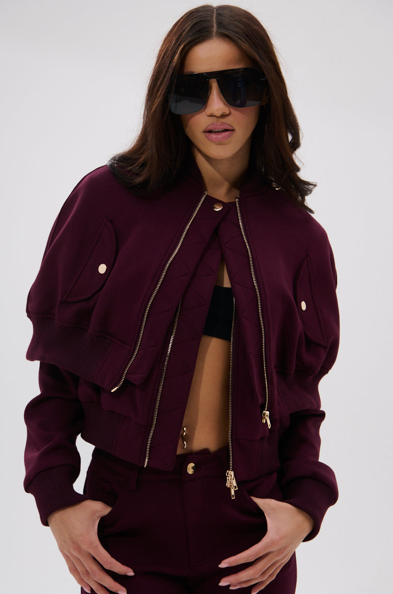 A woman with long brown hair wears large dark sunglasses and the LYNX LAYERED BOMBER JACKET IN BURGUNDY with gold zippers, pairing it with matching high-waisted pants. She poses with one hand in her pocket against a plain light background.