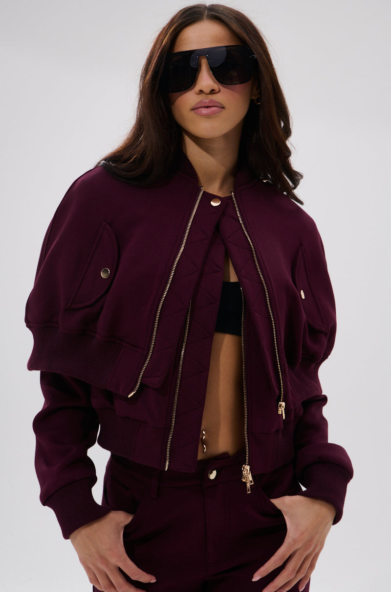 A person confidently poses with hands in pockets against a plain light background, wearing the LYNX LAYERED BOMBER JACKET IN BURGUNDY, styled with large black sunglasses and matching pants featuring gold zippers.