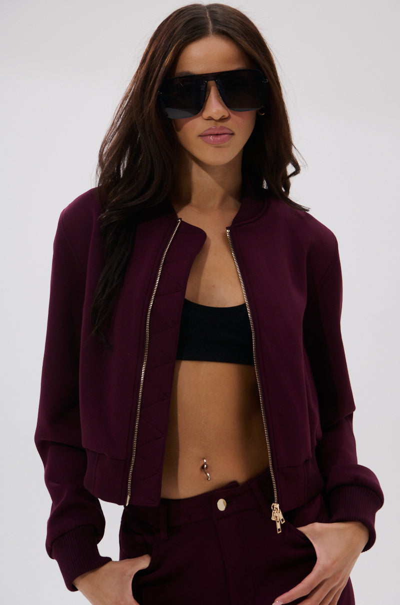 A woman with long dark hair stands against a plain background, hands in her pockets, wearing large black sunglasses and the LYNX LAYERED BOMBER JACKET IN BURGUNDY over a black crop top and maroon pants. Her belly button piercing is visible.