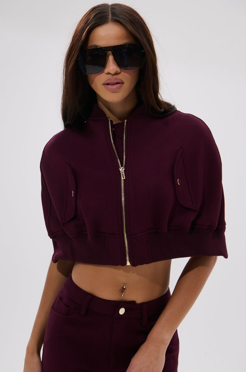 A woman stands against a plain light background wearing the LYNX LAYERED BOMBER JACKET IN BURGUNDY with matching pants and large black sunglasses.