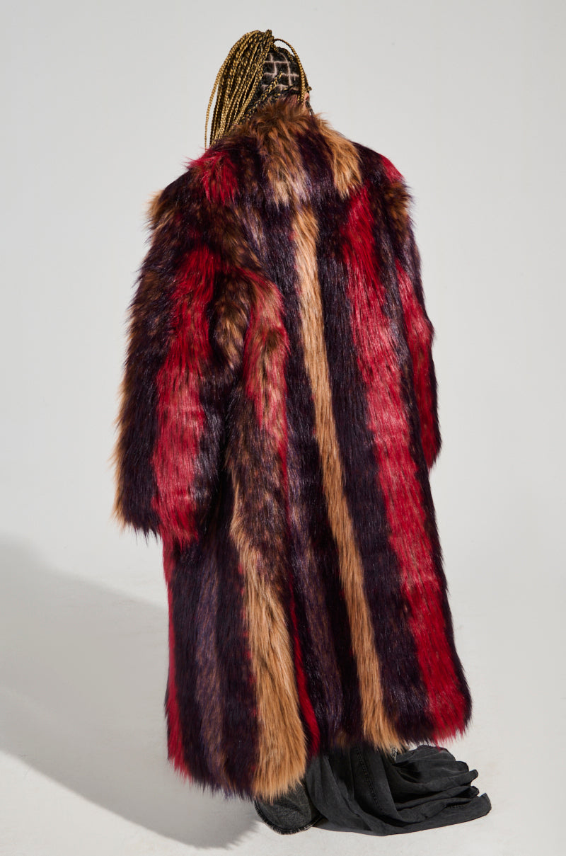 With their back turned, an individual with braided hair highlights an oversized silhouette draped in the SUNDAY STRIPED FUR COAT. The vibrant faux fur coat features striking red, brown, and black stripes that appear luxuriously plush against the light background.