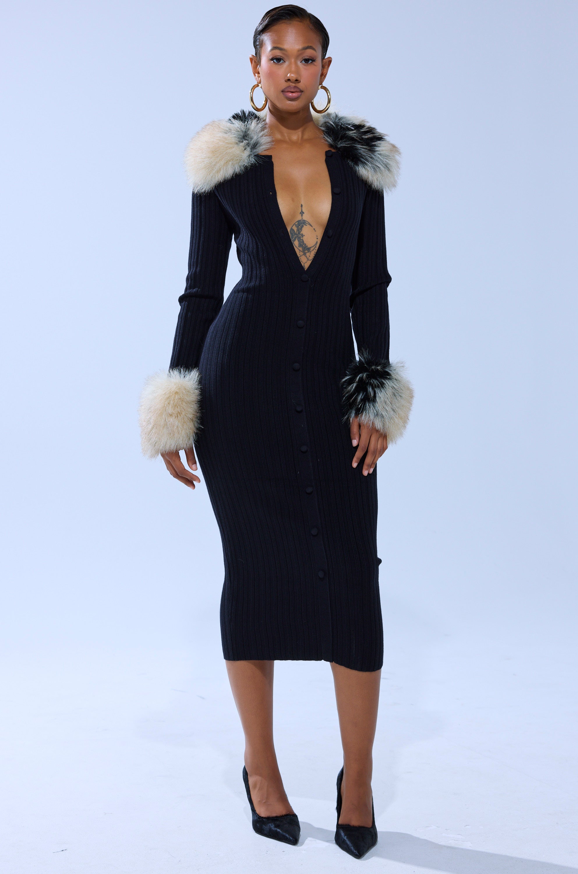 A woman stands confidently in the MICHELLE KNIT MIDI DRESS WITH FUR ACCENTS, featuring a ribbed knit design, deep V-neckline, front buttons, and faux fur on the cuffs and collar. She pairs it with hoop earrings and black heels against a light background.