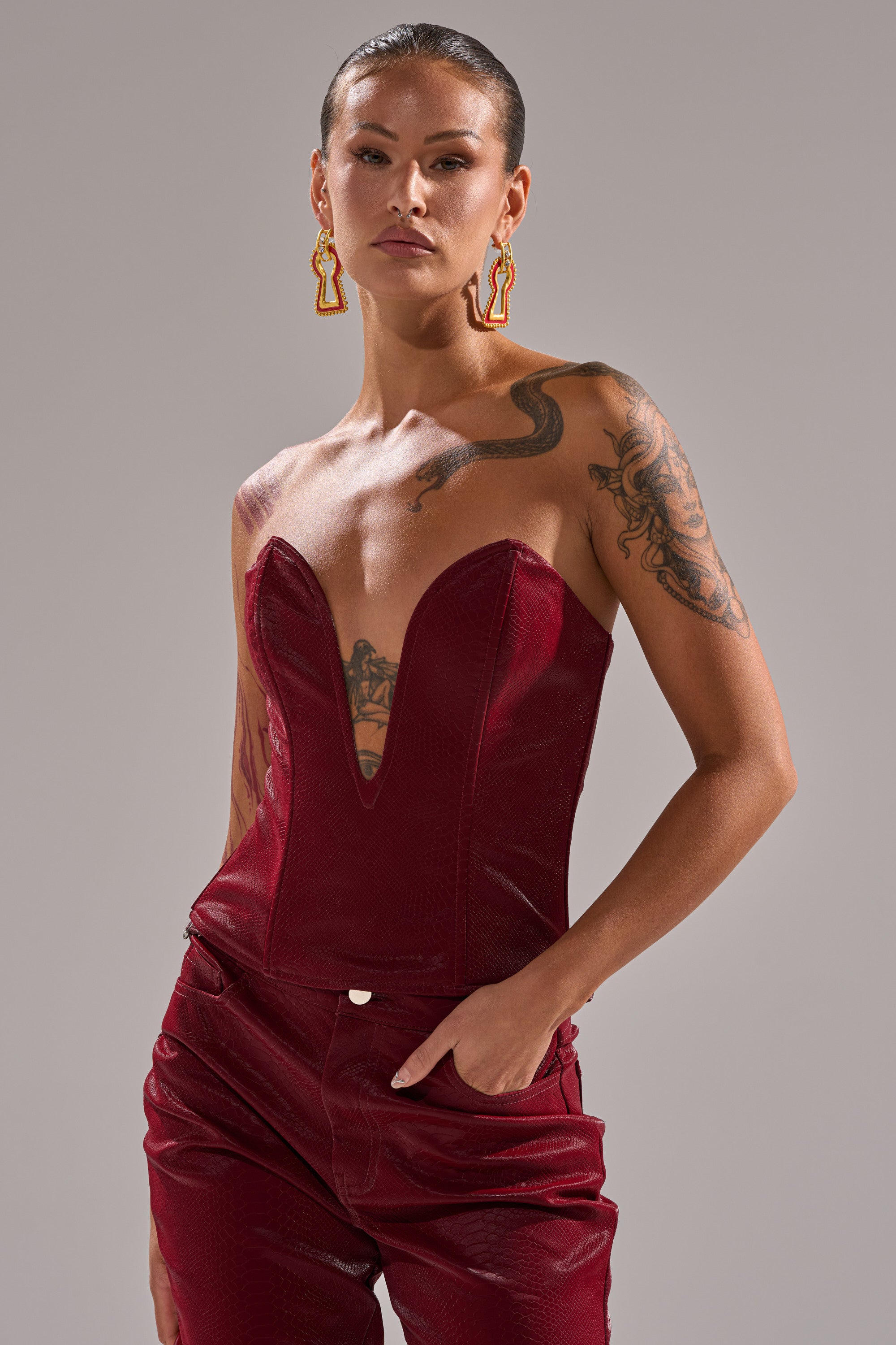 A woman with tattoos wears the TOO HOT FOR YOU FAUX LEATHER CORSET with matching pants, styled with slicked-back hair and statement earrings, posing confidently against a plain gray background.