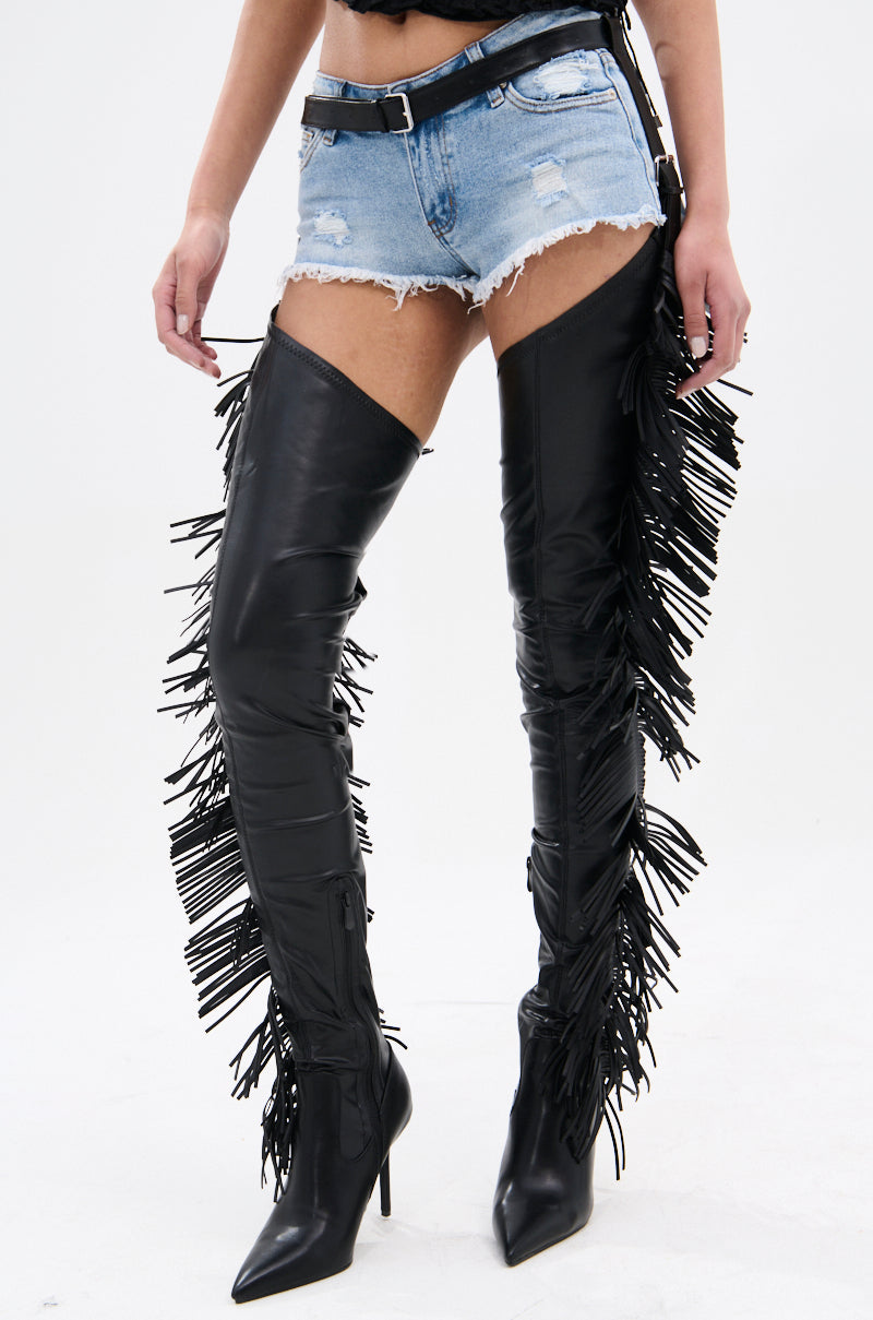 Someone is wearing the AZALEA WANG KARTICE BLACK STILETTO WESTERN CHAP BOOTS, featuring black thigh-high styling, fringe details, and pointed toes. They're paired with blue denim shorts and a black belt, with the faux leather boots contrasting against a plain white background.