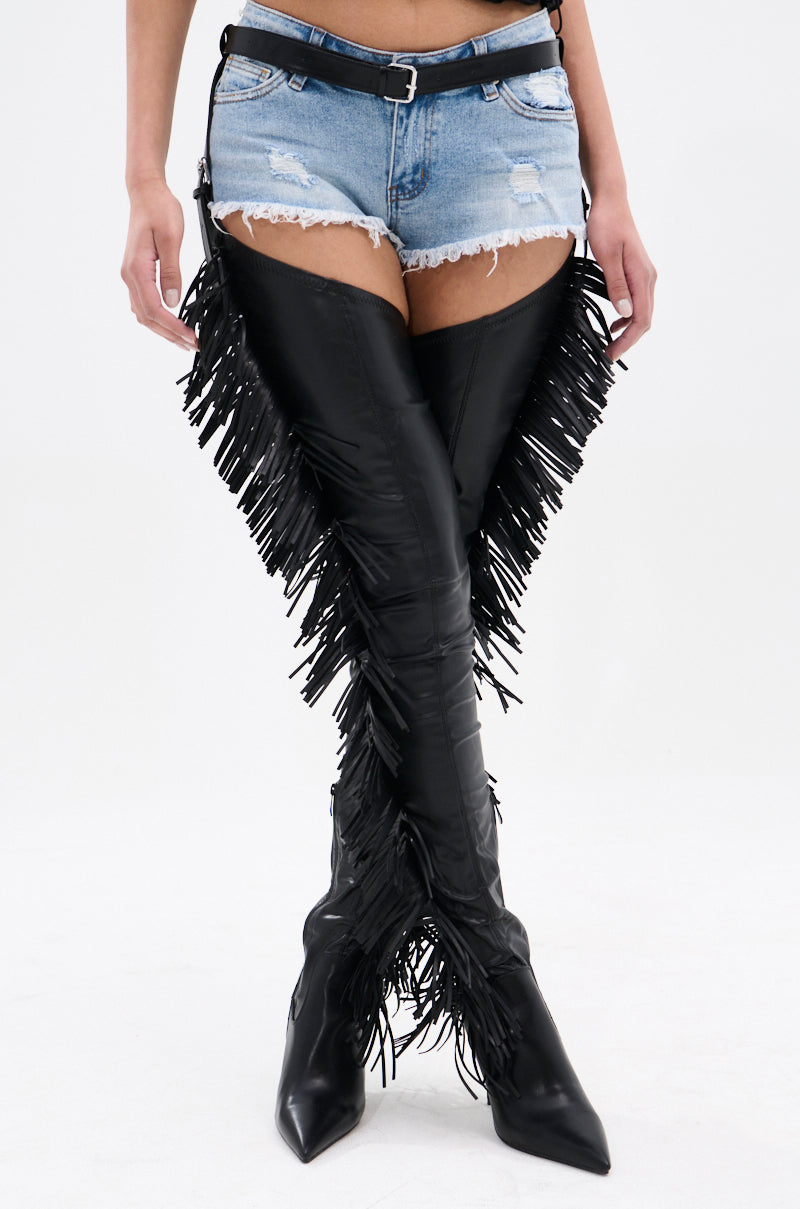 Against a plain background, a person dons distressed denim shorts and the AZALEA WANG KARTICE BLACK STILETTO WESTERN CHAP BOOT with fringe detailing, exuding a bold, edgy vibe.