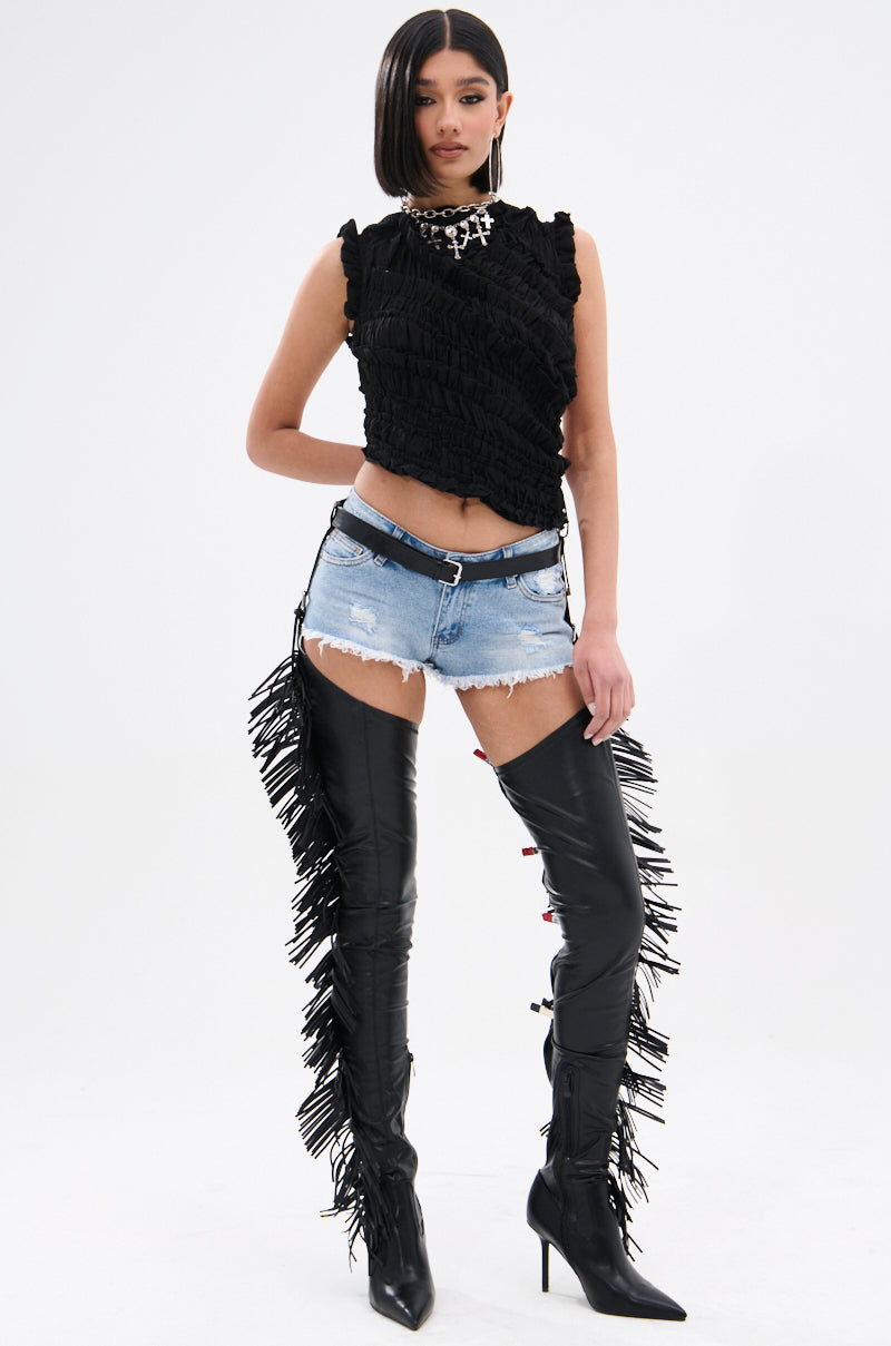 Against a plain background, a person confidently wears a black sleeveless top, denim shorts with a black belt, and AZALEA WANG KARTICE BLACK STILETTO WESTERN CHAP BOOTS adorned with fringe.