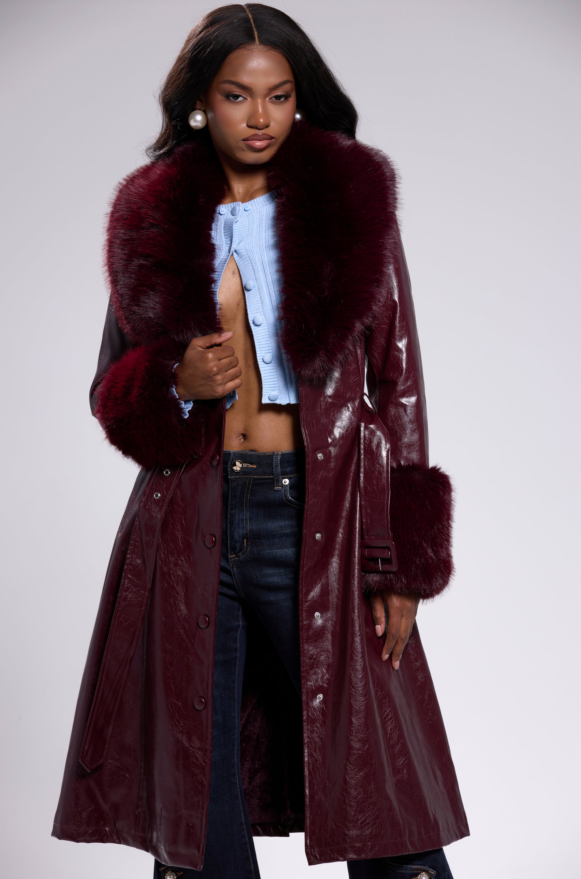 A woman stands confidently against a plain background, wearing the KAYA FUR TRIM TRENCH IN BURGUNDY—a glossy vegan leather coat with large faux fur trim—styled over a light blue cropped top and dark jeans.