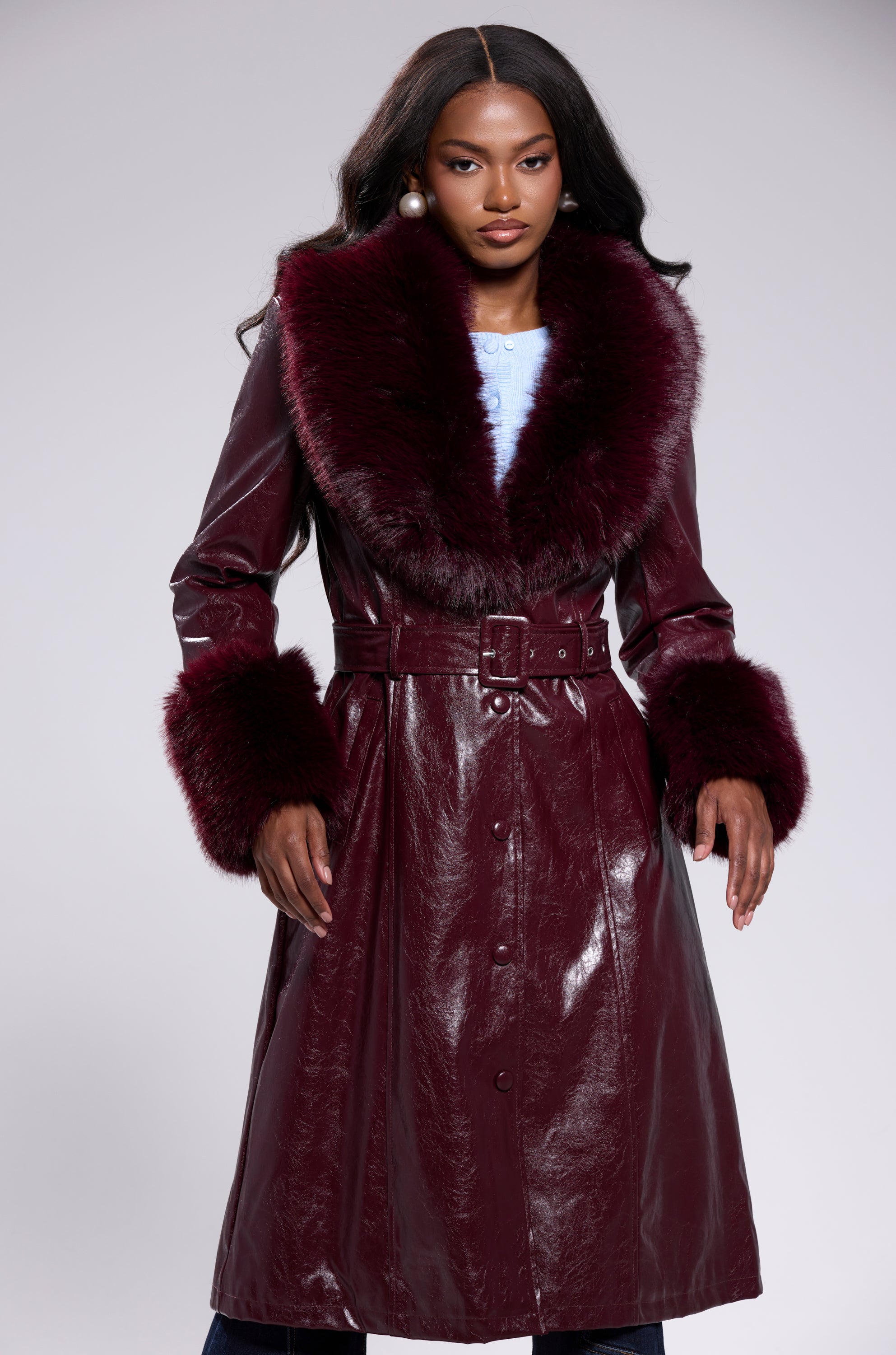 A woman stands confidently against a light background wearing the KAYA FUR TRIM TRENCH IN BURGUNDY, a shiny vegan leather coat with wide faux fur collar and cuffs, belted at the waist. She has long wavy hair and pearl earrings.