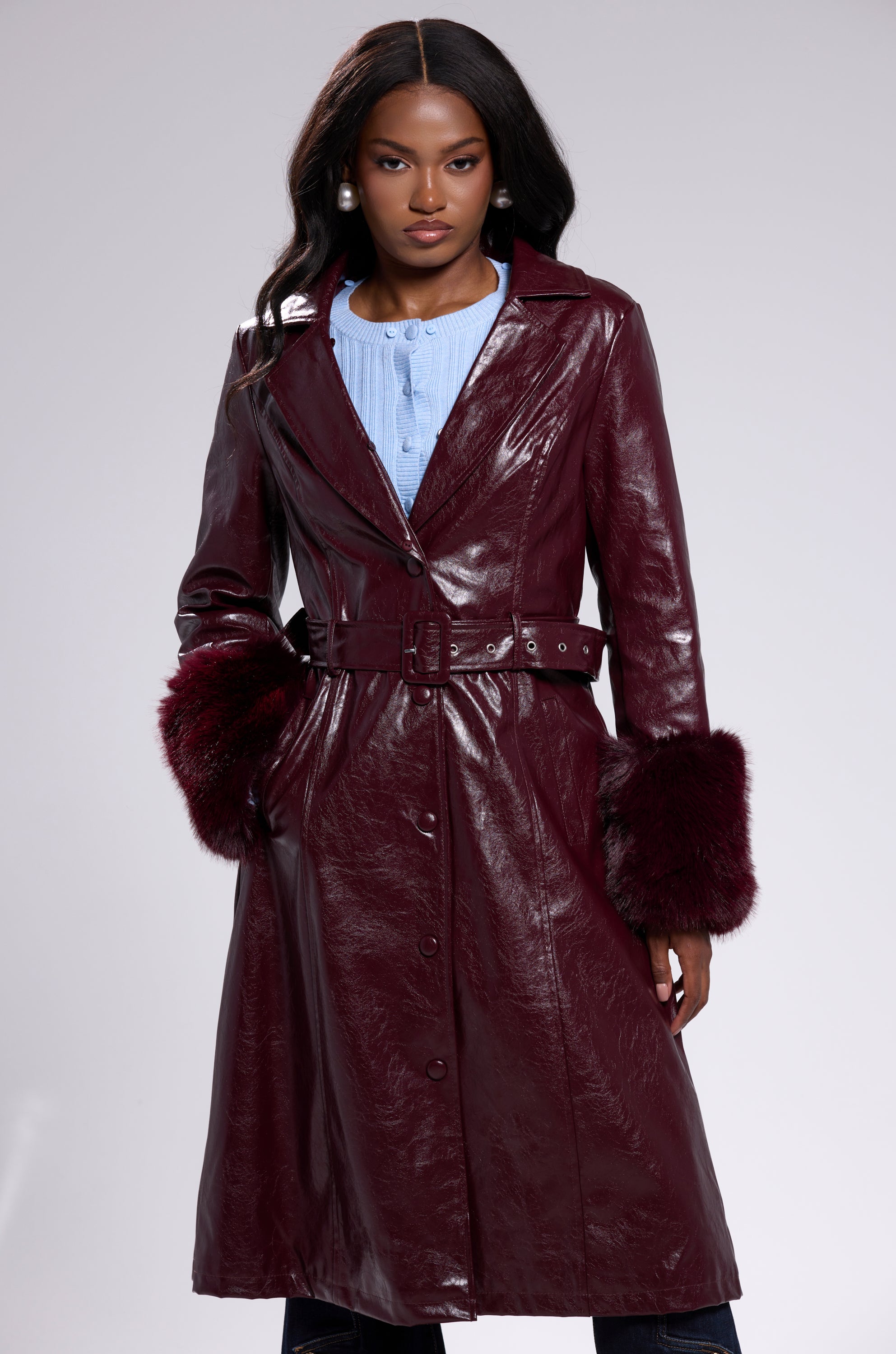 A woman wears the KAYA FUR TRIM TRENCH IN BURGUNDY—a long, belted trench with faux fur trim—over a light blue buttoned shirt, standing with her hands in her pockets against a plain background.